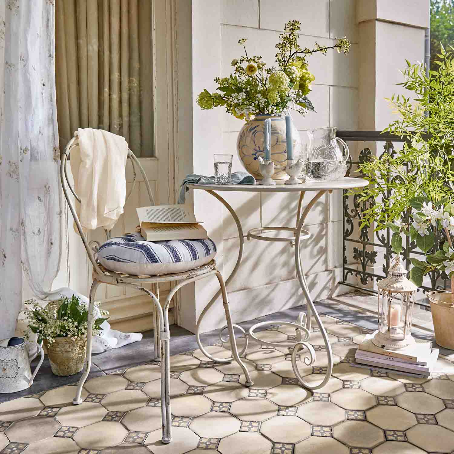 Mesa y silla metálicas blancas con cojín a rayas en terraza con flores y farol decorativo