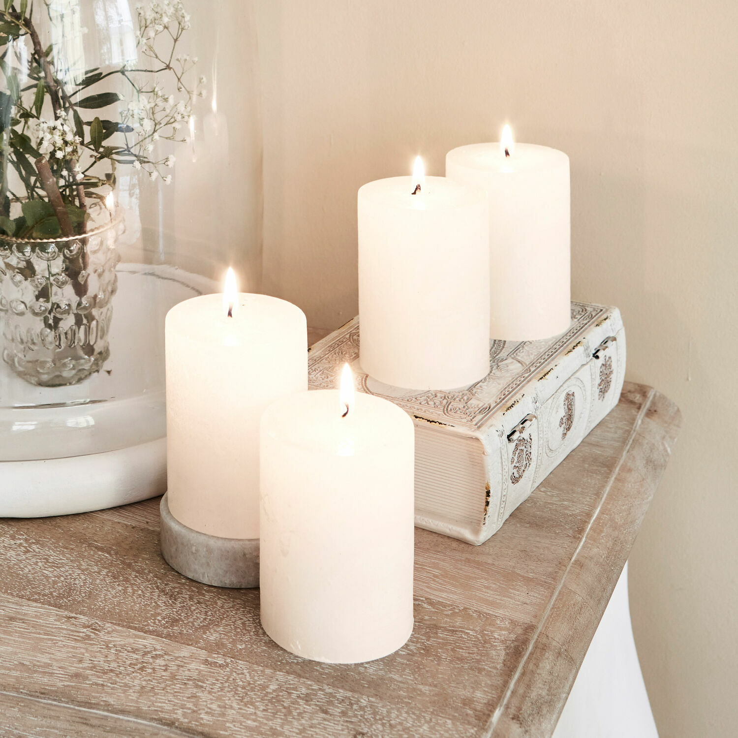Set de 4 velas Virrans Cuatro velas blancas decorativas sobre mesa de madera con libro decorativo y jarrón de cristal