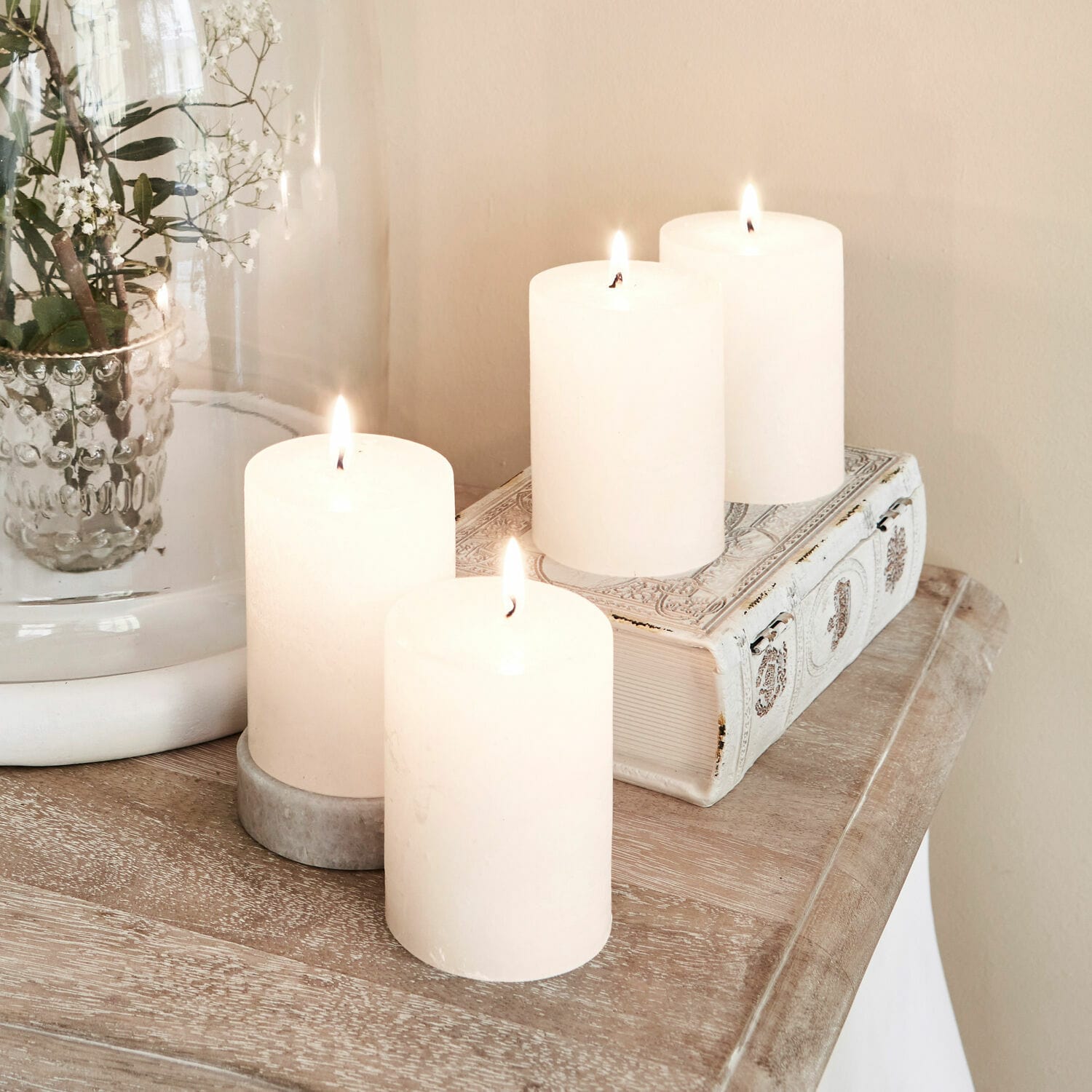 Set de 4 velas Virrans Cuatro velas blancas decorativas sobre mesa de madera con libro decorativo y jarrón de cristal
