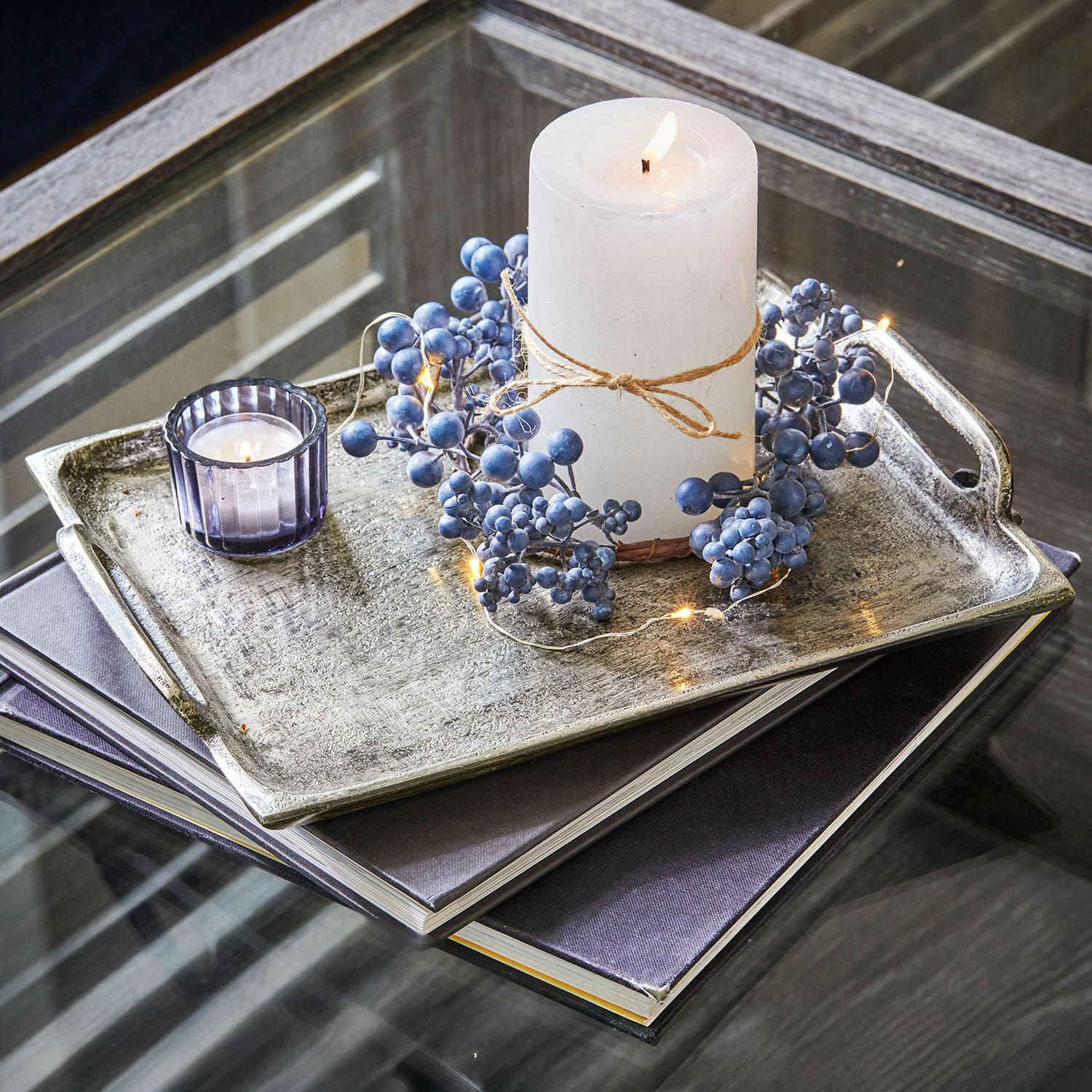 Set de 2 bandejas Branora Vela blanca con cuerda natural y decoración de bayas azules sobre bandeja metálica en mesa de cristal