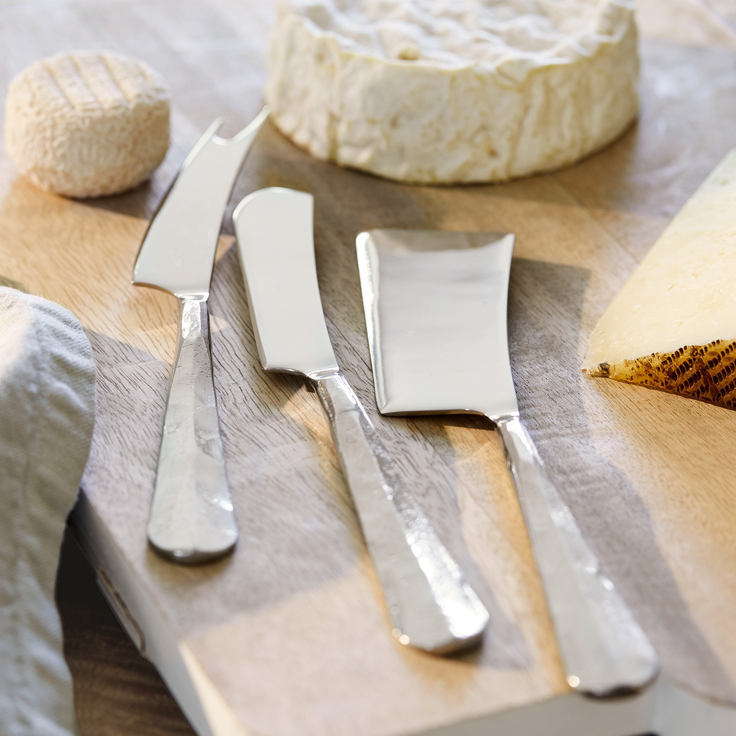 Set de 3 cubiertos para queso Mondevert Conjunto de tres cuchillos para queso de acero inoxidable sobre tabla de madera con quesos variados