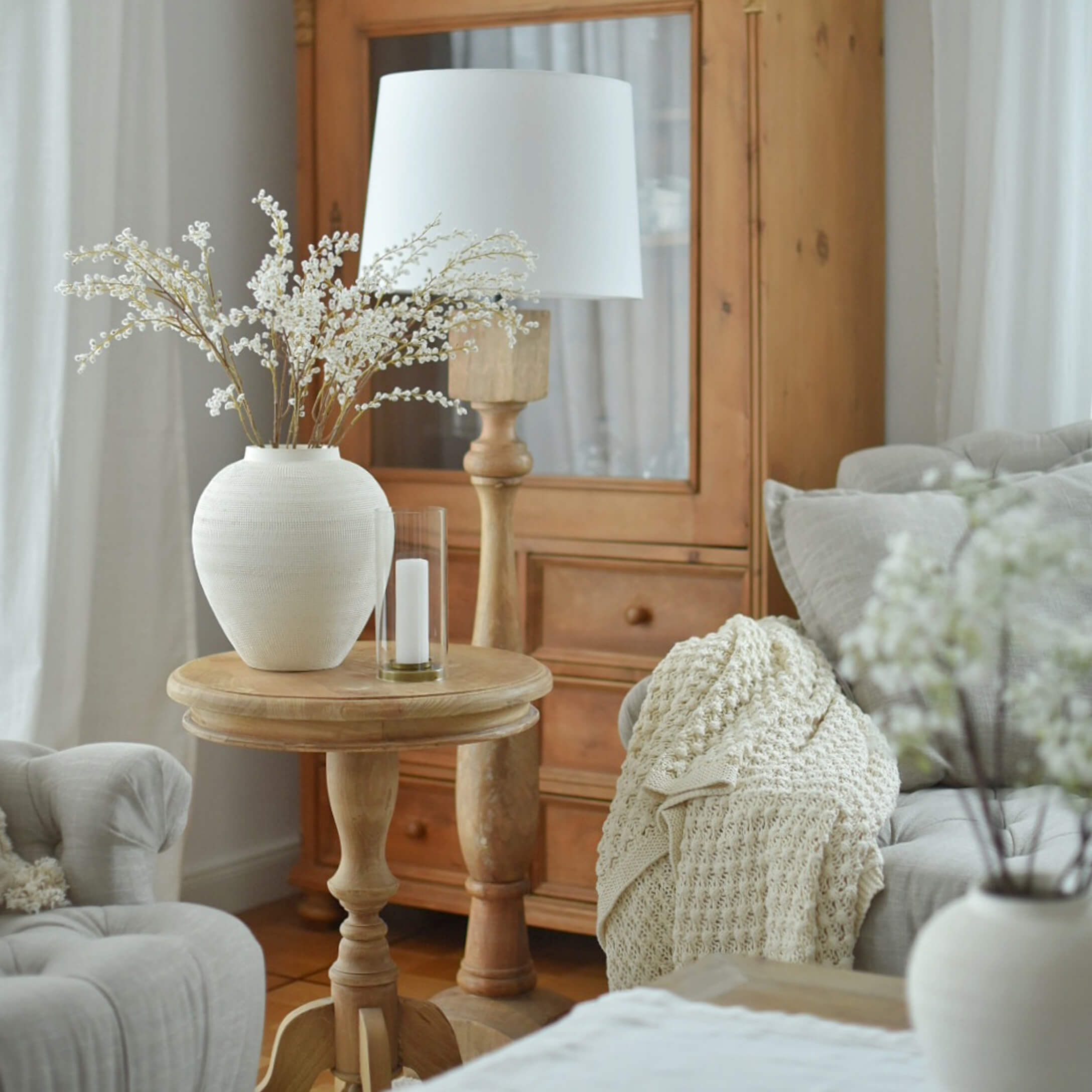 Mesa de madera clara con jarrón de cerámica blanca y lámpara en salón acogedor con manta beige
