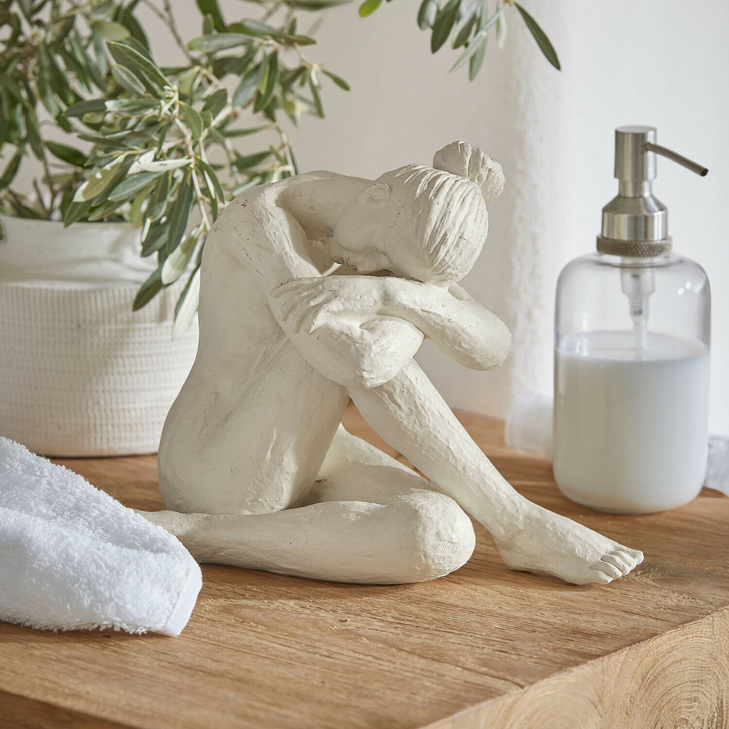 Figura Savigné Escultura blanca de mujer sentada con la cabeza sobre las rodillas dobladas sobre una superficie de madera en baño
