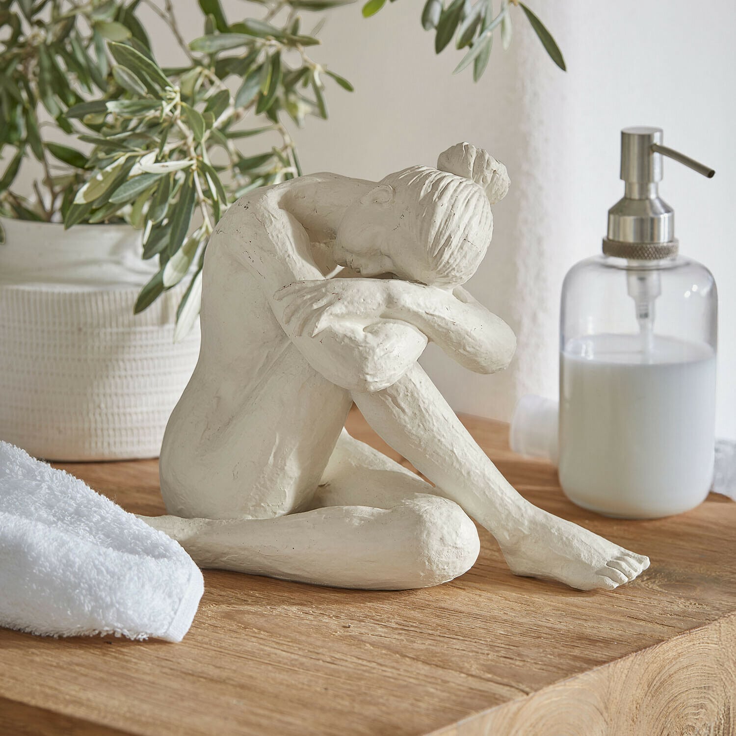 Figura Savigné Escultura blanca de mujer sentada con la cabeza sobre las rodillas dobladas sobre una superficie de madera en baño