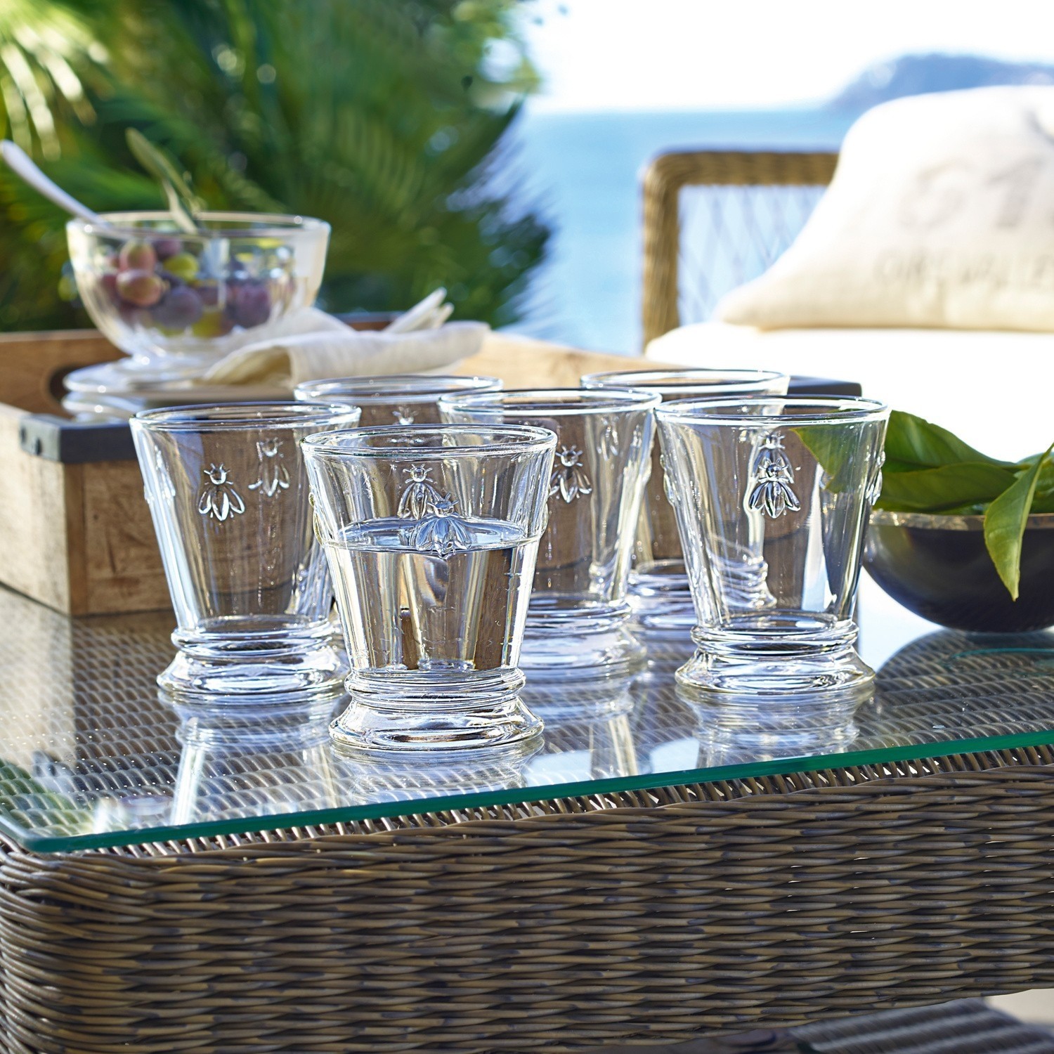 Set de 6 vasos de agua Abeille Juego de seis vasos de vidrio transparente con diseño de abeja sobre mesa de cristal al aire libre