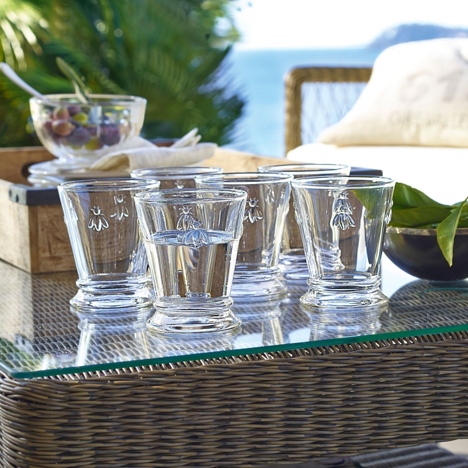 Set de 6 vasos de agua Abeille Juego de seis vasos de vidrio transparente con diseño de abeja sobre mesa de cristal al aire libre