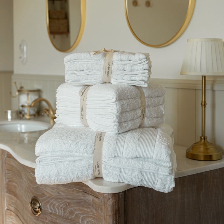Conjunto de toallas blancas de algodón apiladas sobre una cómoda de madera en un baño con espejo y lámpara