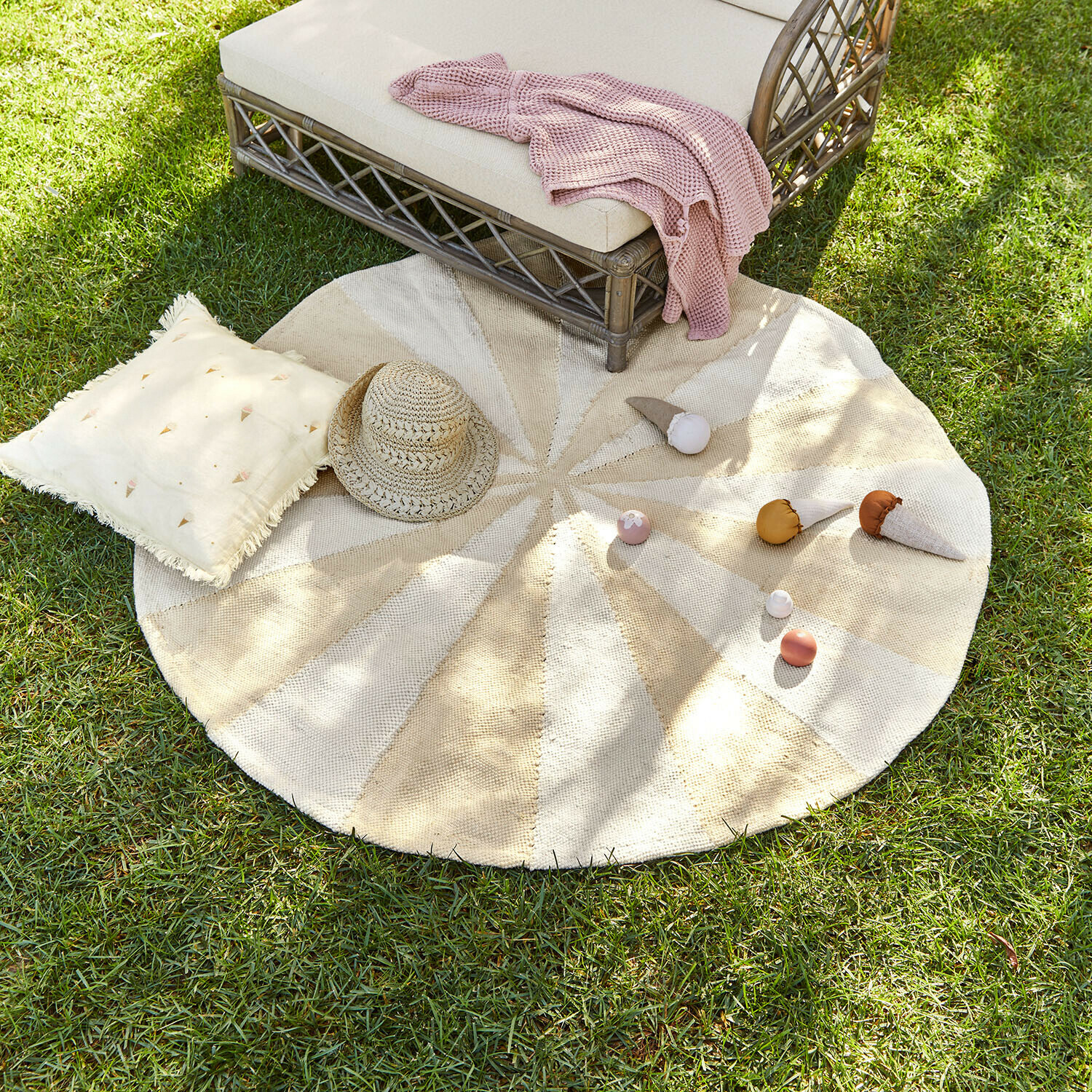 Alfombra redonda de material natural en tonos beige y crema sobre césped con cojín y sombrero de paja