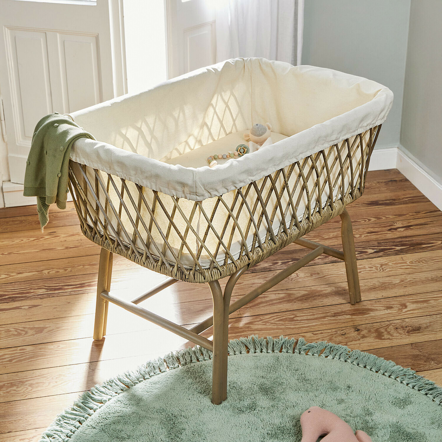 Cuna de mimbre con forro crema y manta verde sobre patas de madera en habitación infantil