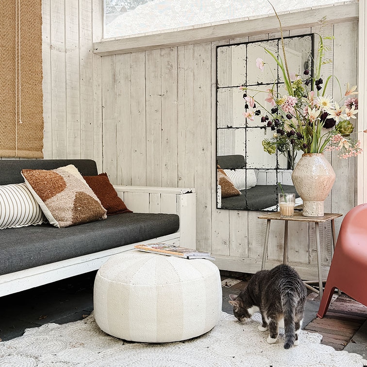 Salón acogedor con pared de madera blanca, sofá gris, puf blanco y decoración floral sobre mesa de madera