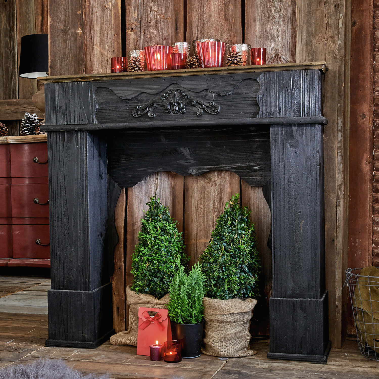 Consola de chimenea Margaux Chimenea decorativa de madera negra con plantas verdes y velas rojas en interior rústico