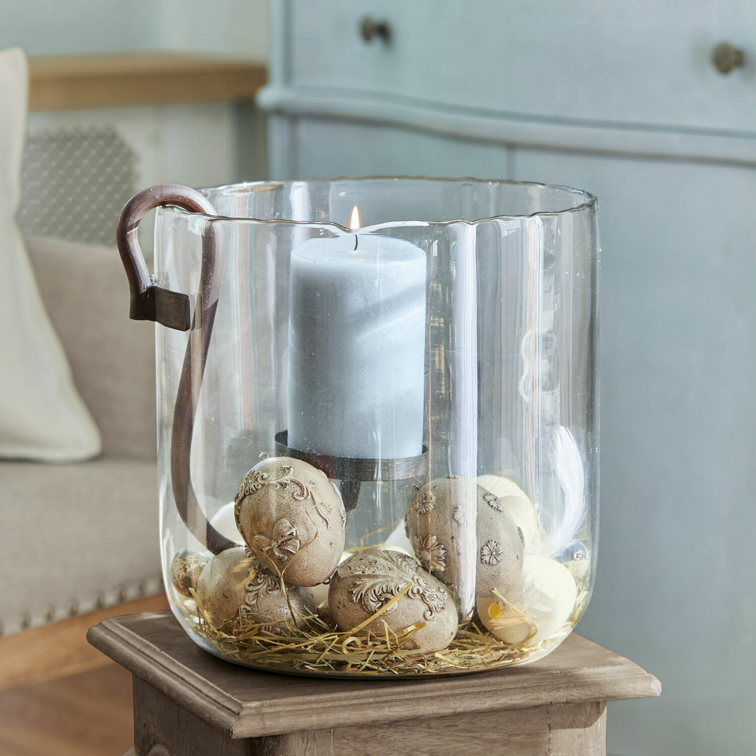 Portavelas de vidrio con asa, vela gris y piedras decorativas sobre paja para decoración interior