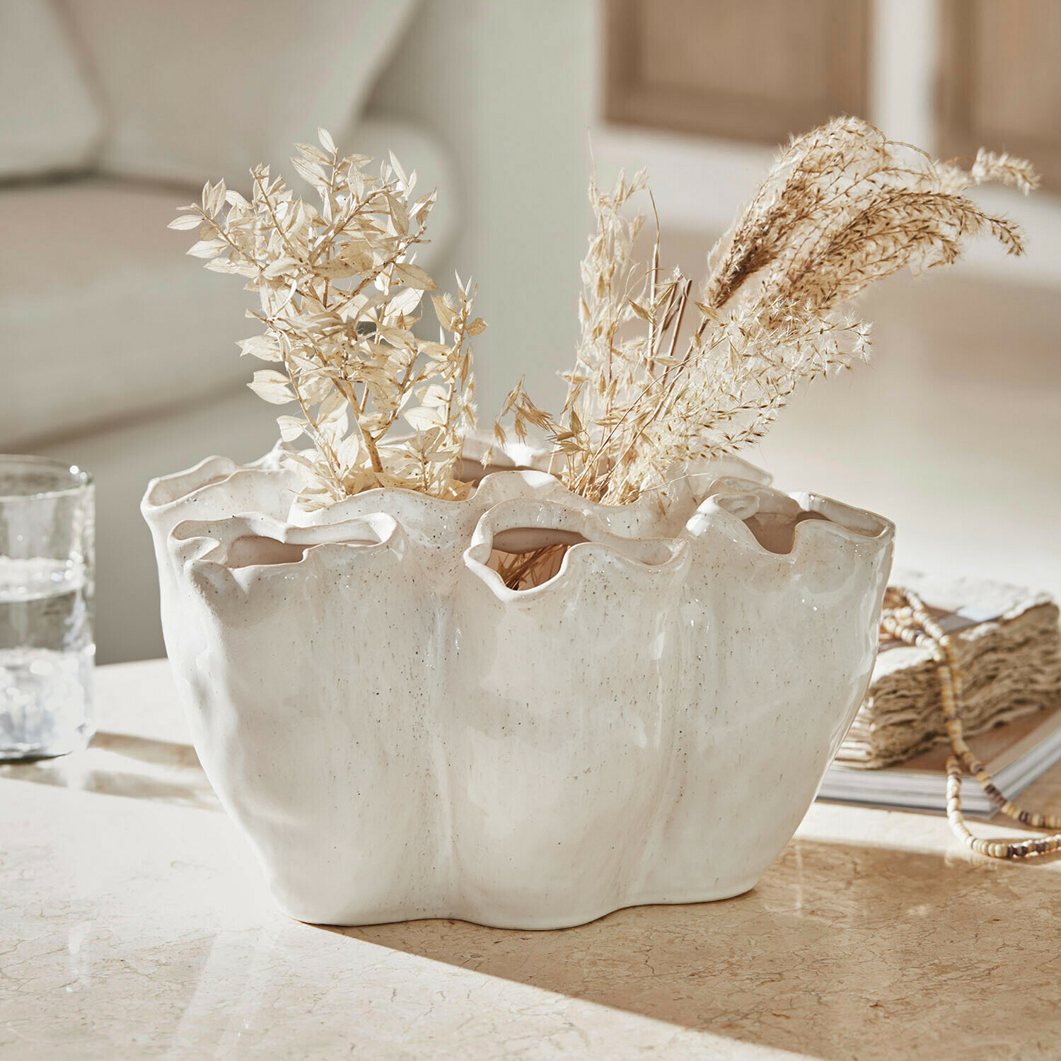 Jarrón de cerámica blanca con borde ondulado y plantas secas sobre mesa decorativa