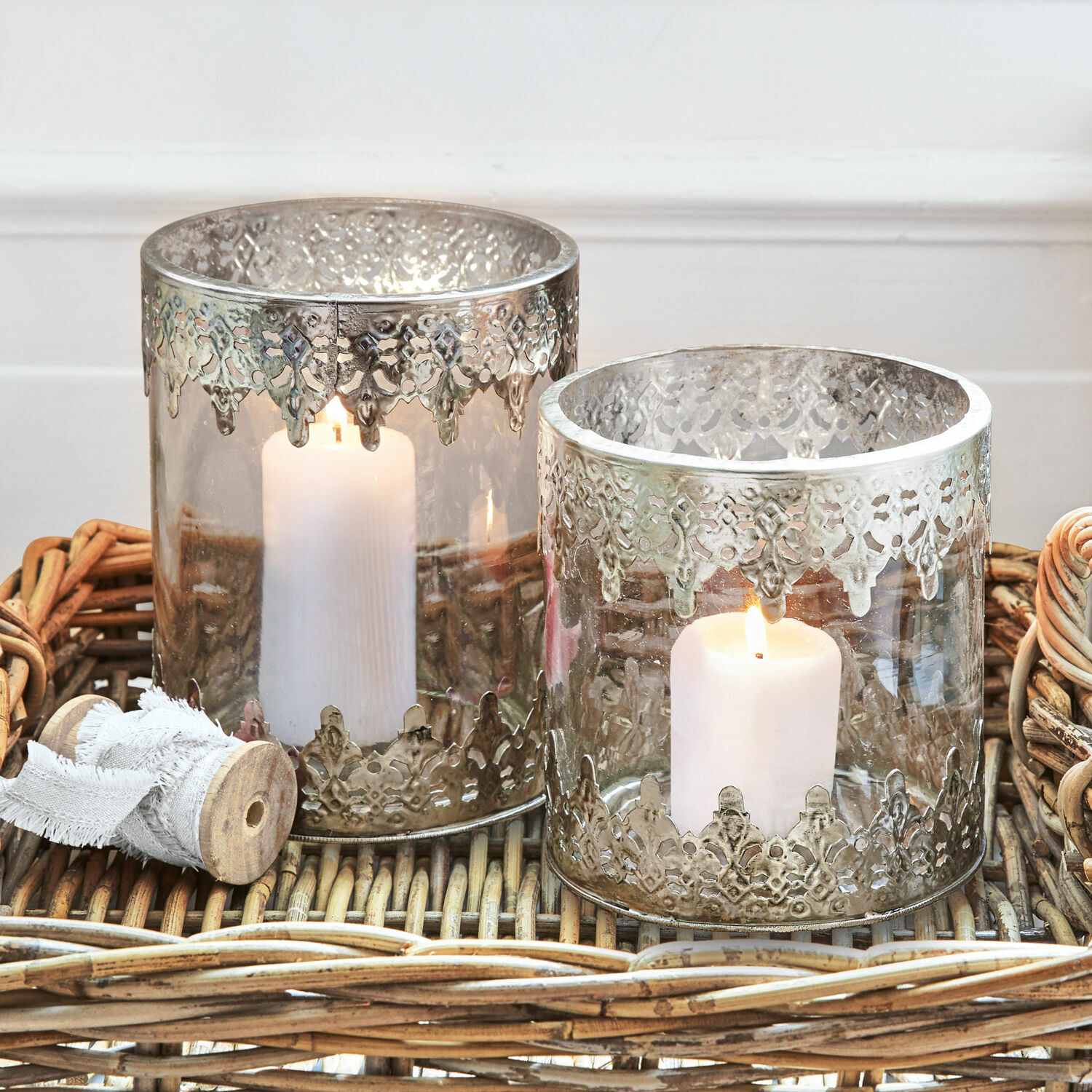Dos portavelas de vidrio con decoración metálica plateada calada sobre bandeja de mimbre
