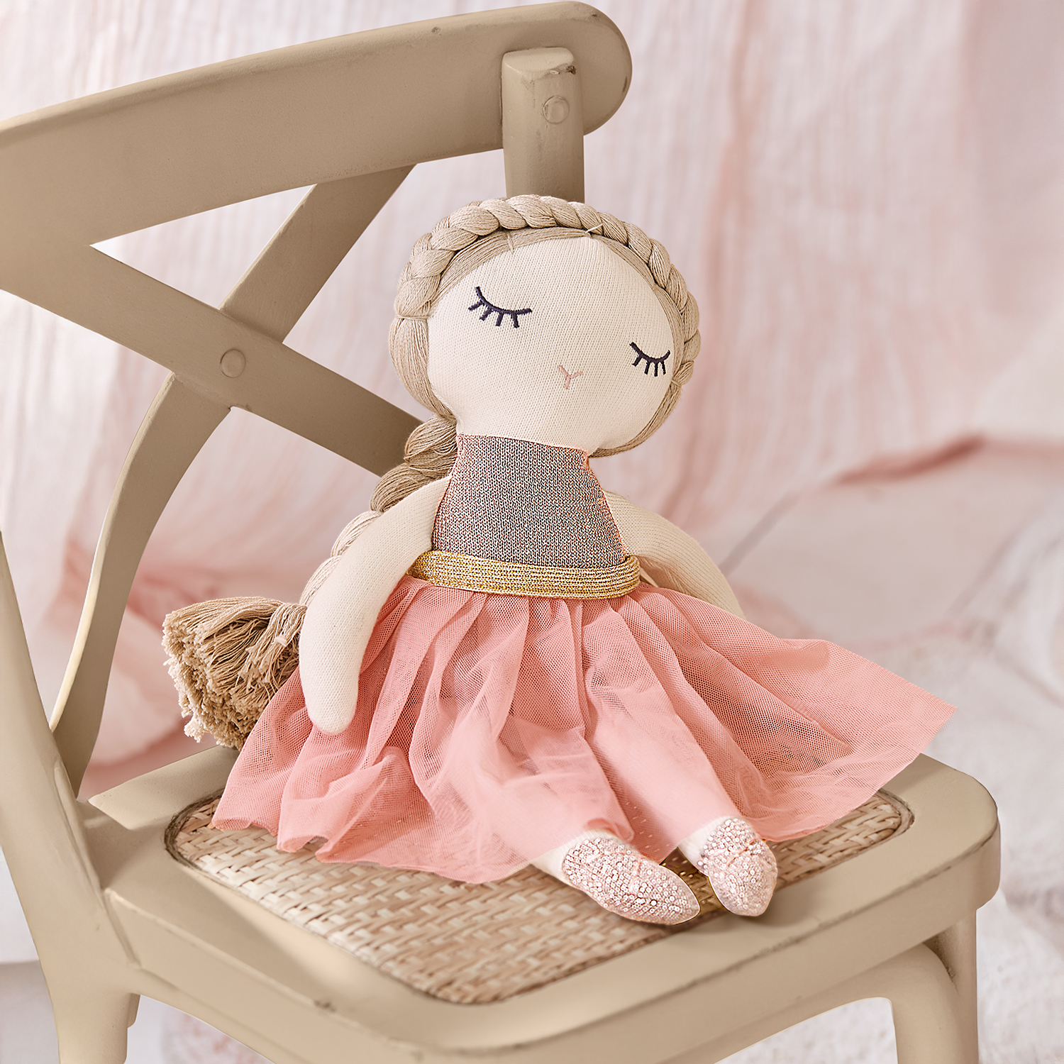 Muñeca de tela con vestido rosa y cinturón dorado sentada en silla beige, juguete infantil