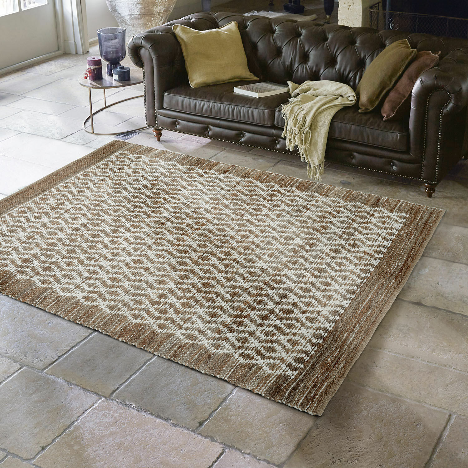 Alfombra reversible Amhara Alfombra marrón y crema con patrón geométrico sobre suelo de piedra frente a sofá de cuero marrón oscuro