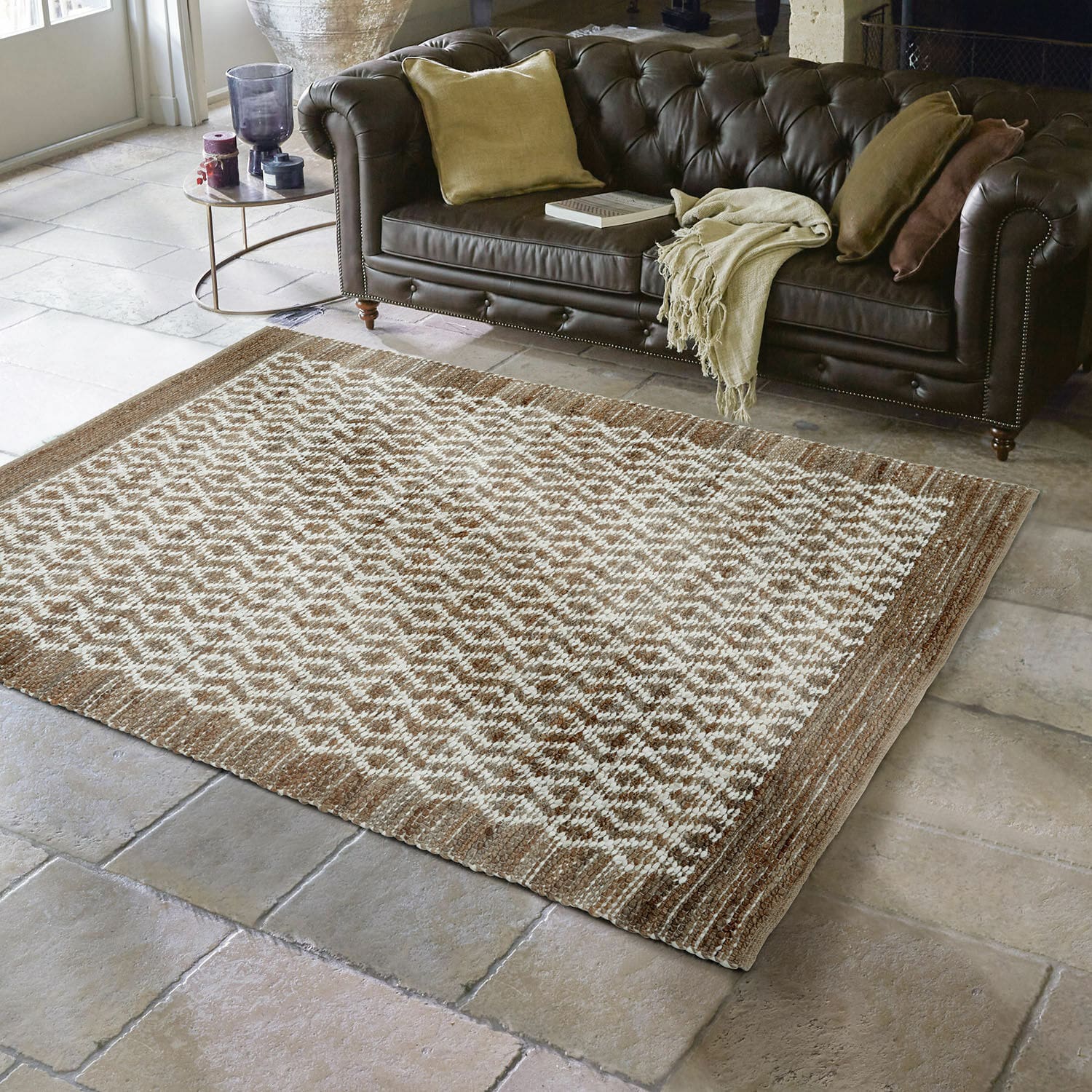 Alfombra reversible Amhara Alfombra marrón y crema con patrón geométrico sobre suelo de piedra frente a sofá de cuero marrón oscuro