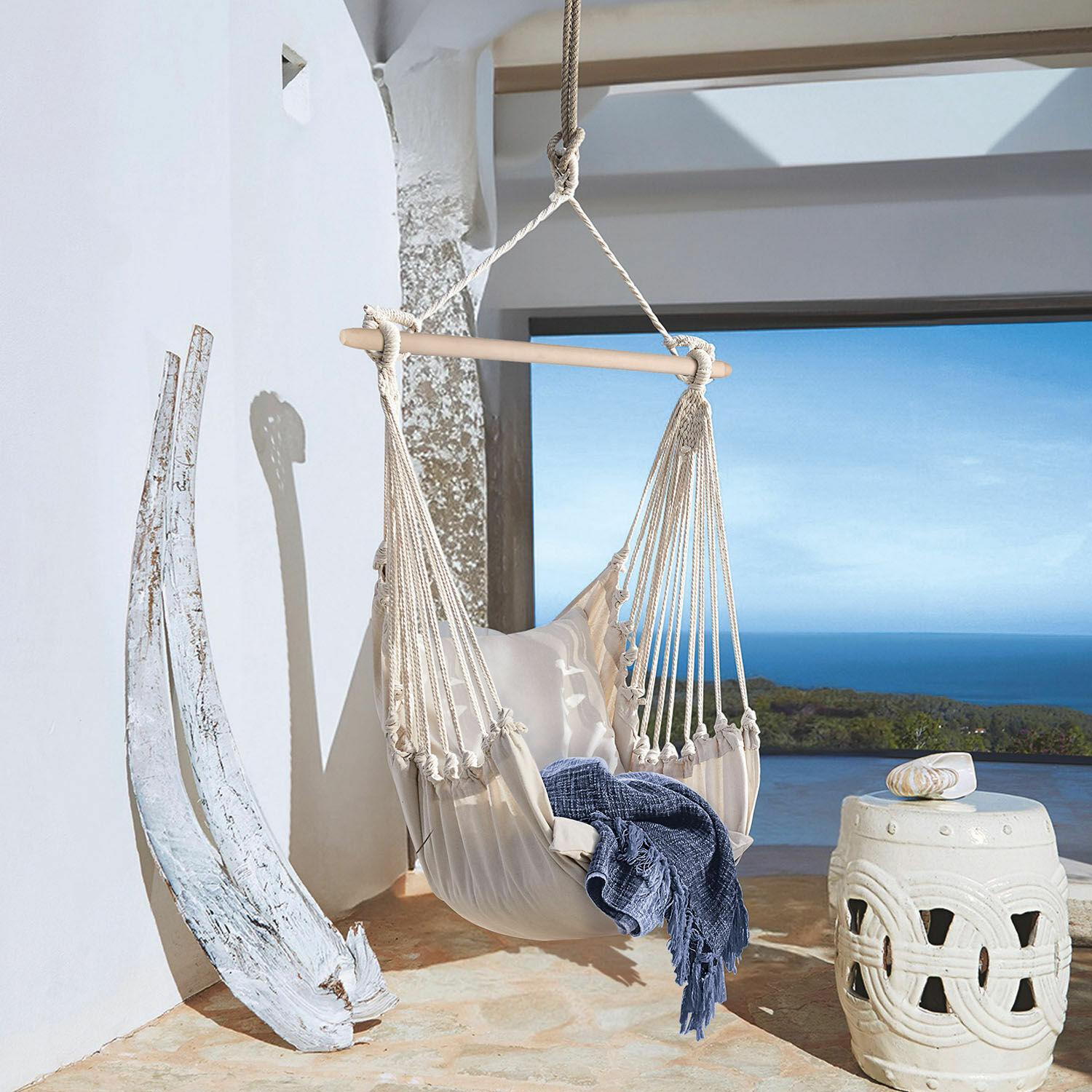 Silla colgante Leano Hamaca beige de algodón con barra de madera y manta azul en terraza con vistas al mar