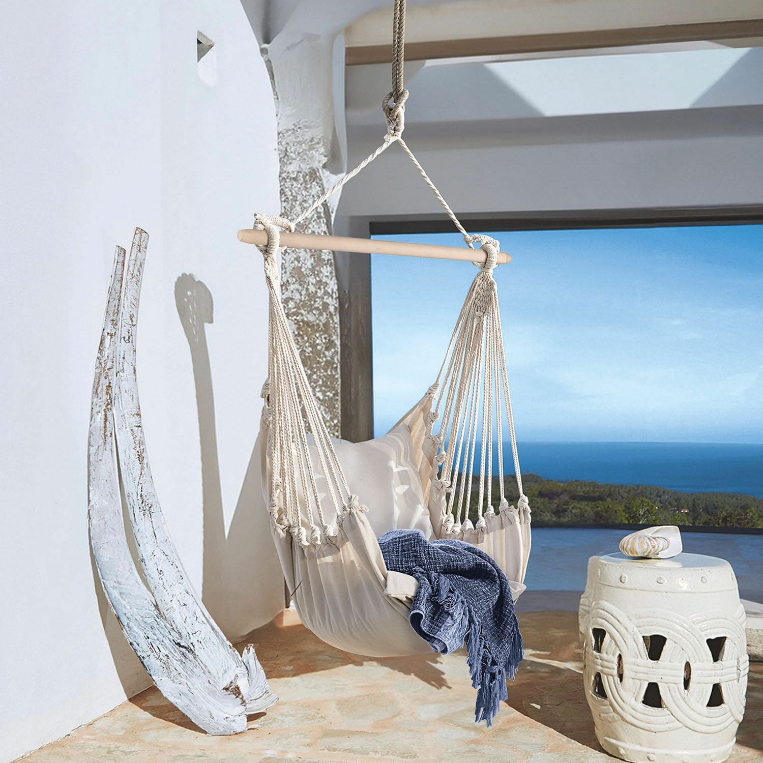 Silla colgante Leano Hamaca beige de algodón con barra de madera y manta azul en terraza con vistas al mar