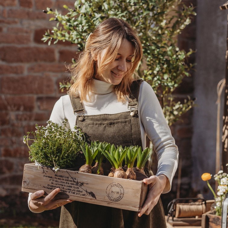 Mujer con peto de pana marrón sosteniendo caja de madera con plantas verdes en jardín