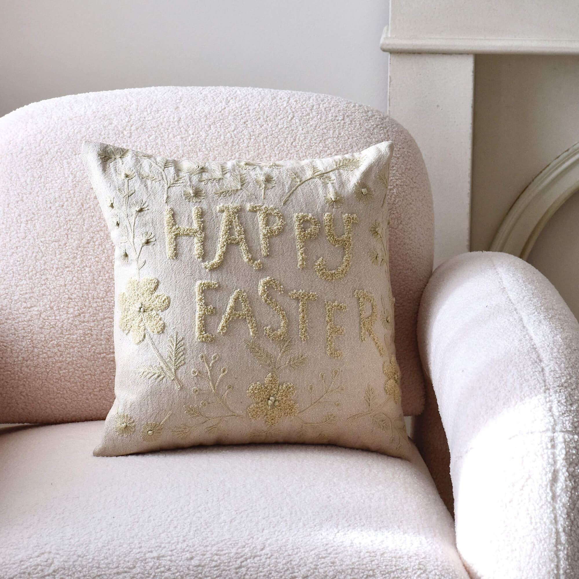 Cojín decorativo beige con bordado "Happy Easter" sobre sillón claro, diseño pastel de Pascua