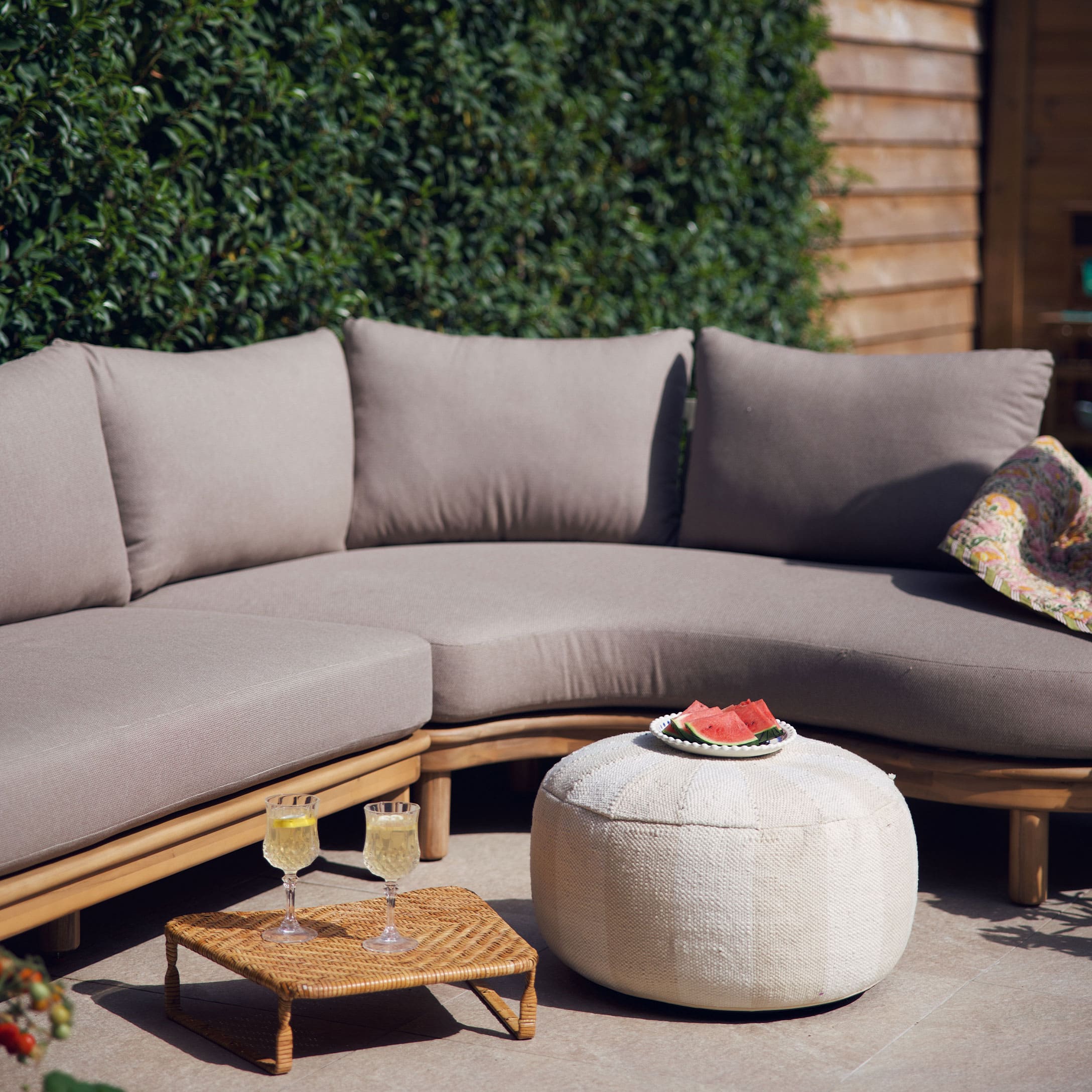 Sofá beige con cojines sobre estructura de madera y puf blanco en terraza decorada para verano