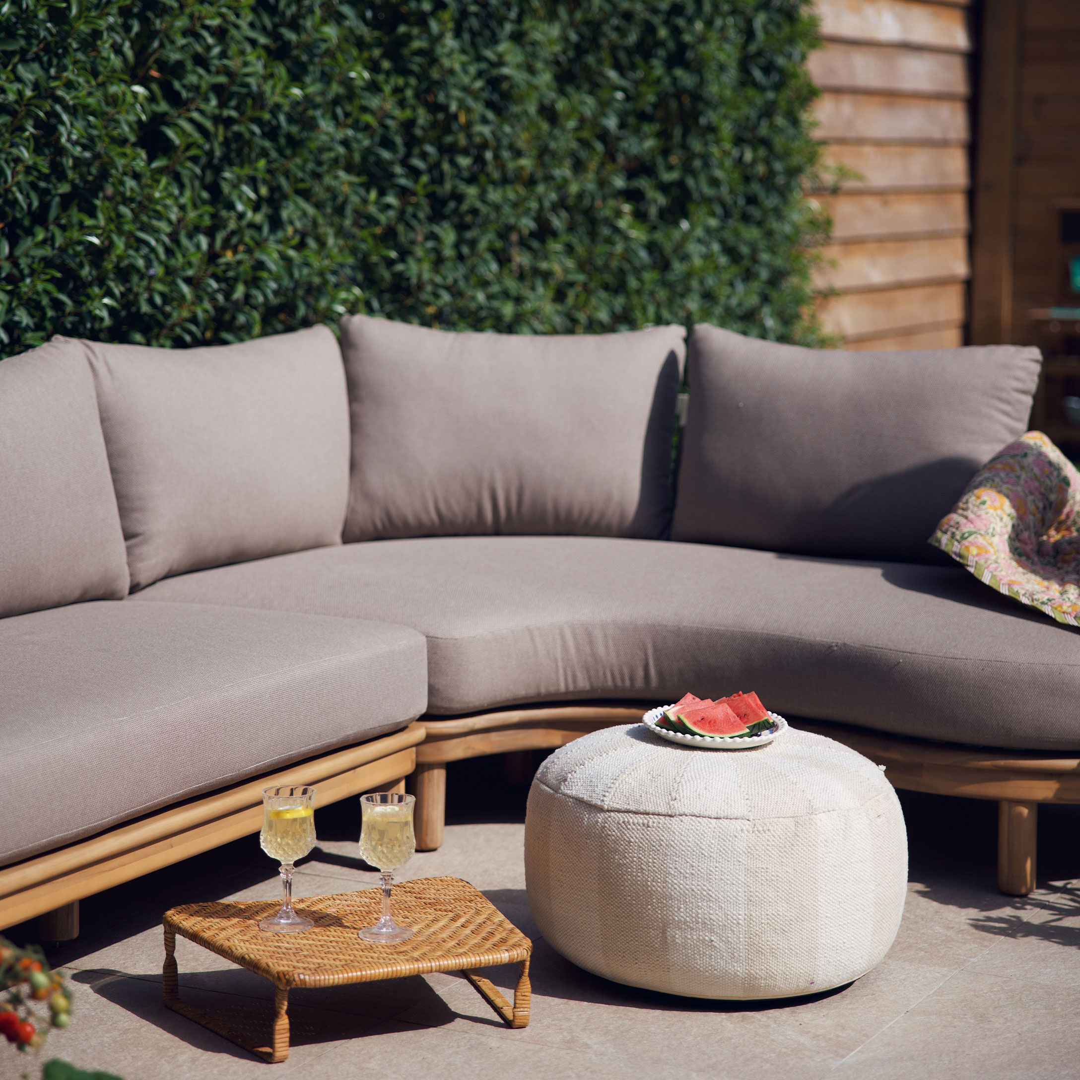 Sofá beige con cojines sobre estructura de madera y puf blanco en terraza decorada para verano
