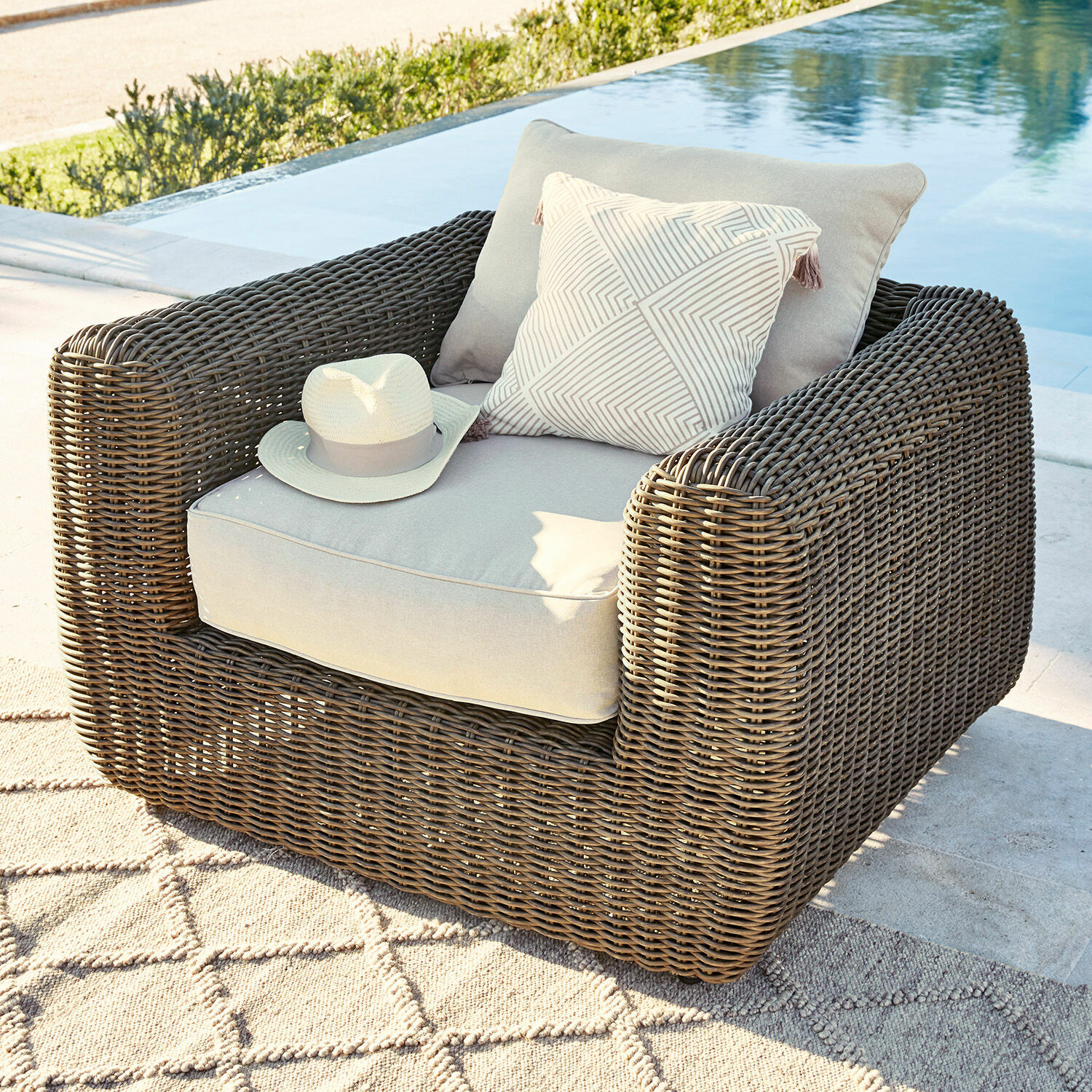 Sillón de jardín de ratán marrón con cojines gris claro y cojín decorativo junto a la piscina