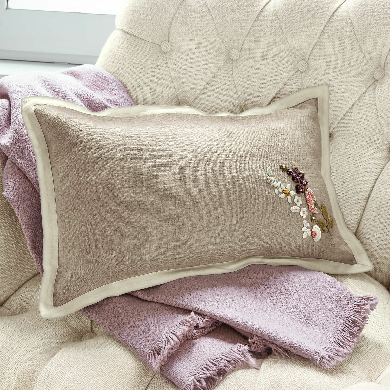 Funda de cojín Tavaline Cojín beige de lino con bordado floral sobre manta rosa claro en sillón, decoración hogareña