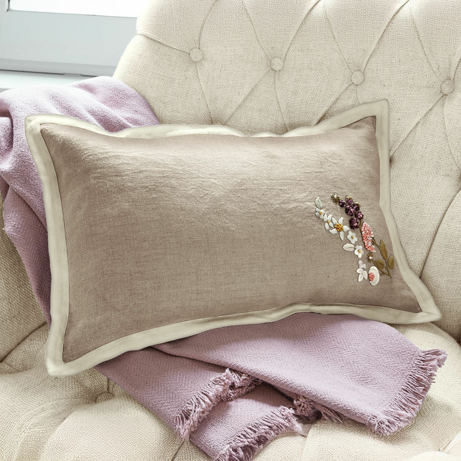 Funda de cojín Tavaline Cojín beige de lino con bordado floral sobre manta rosa claro en sillón, decoración hogareña