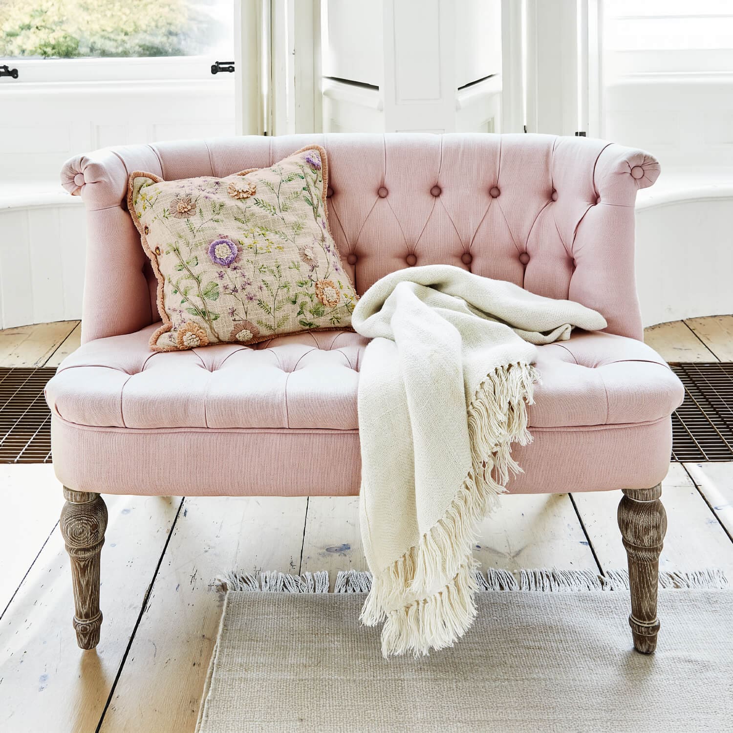 Sofá tapizado rosa con patas de madera, cojín decorativo y manta crema sobre suelo claro