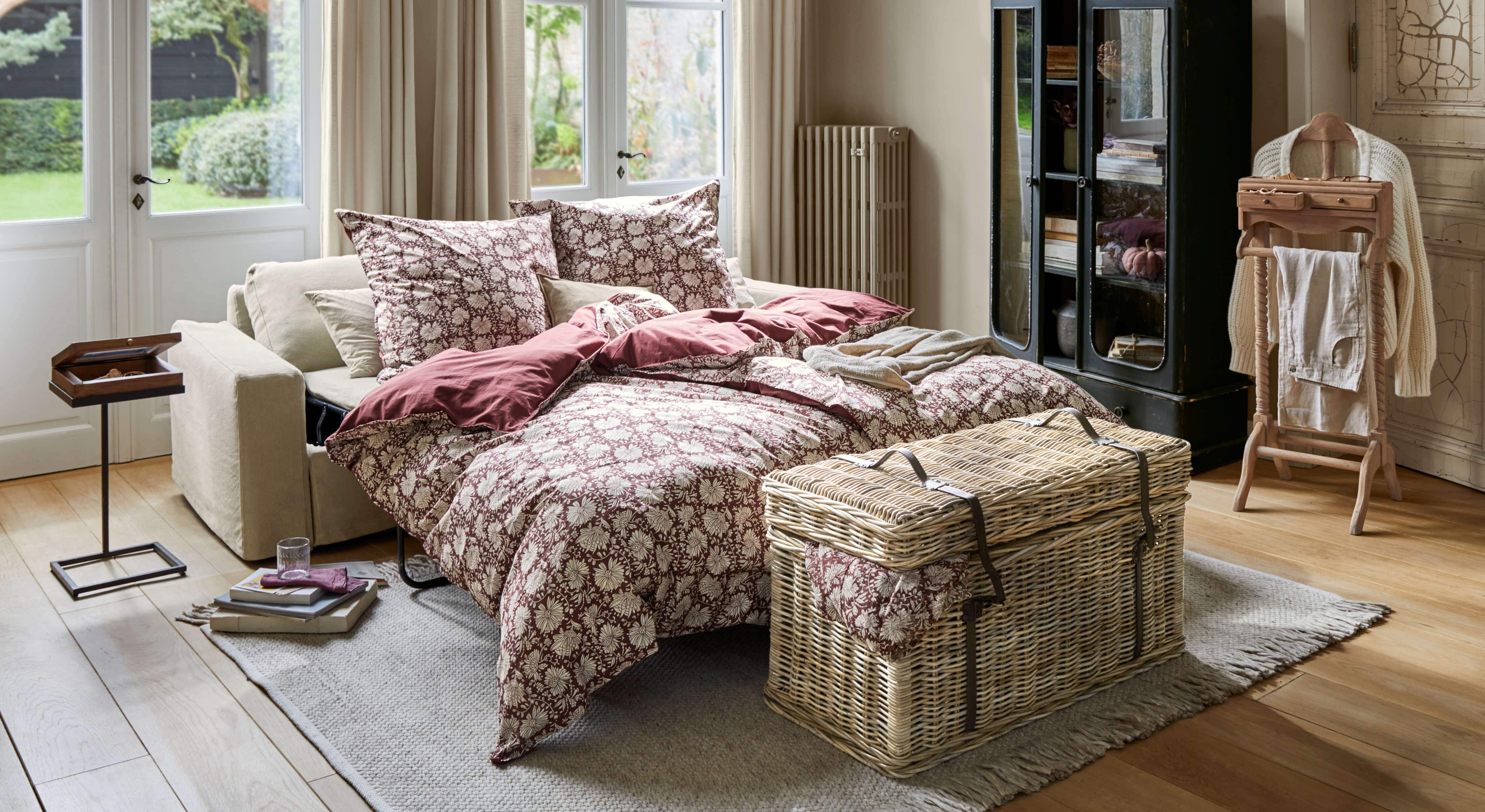 Sofá cama beige con ropa de cama burdeos con estampado floral en salón luminoso con suelo de madera