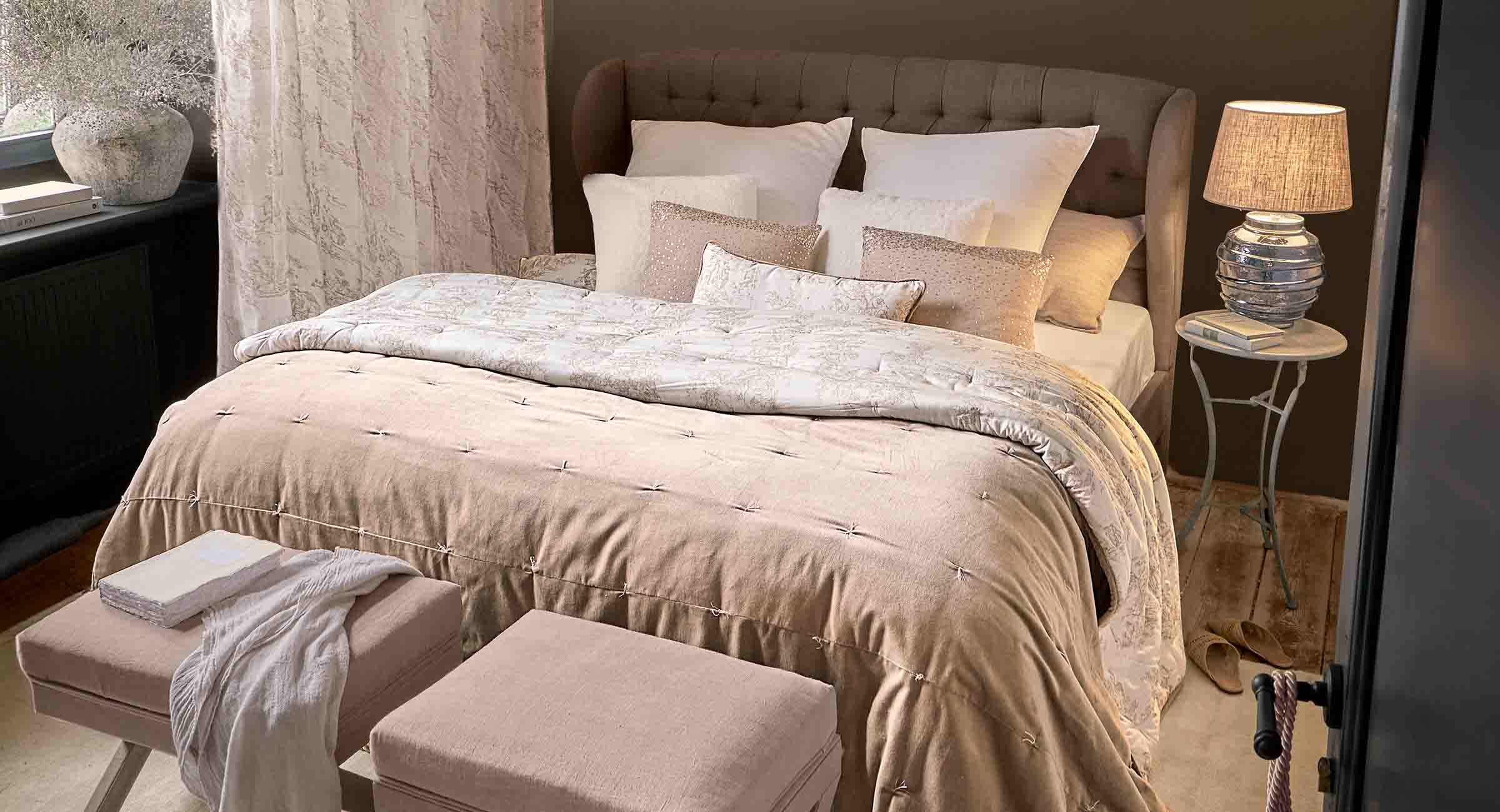 Dormitorio acogedor con colcha beige y cojines sobre cama tapizada, lámpara sobre mesita de noche