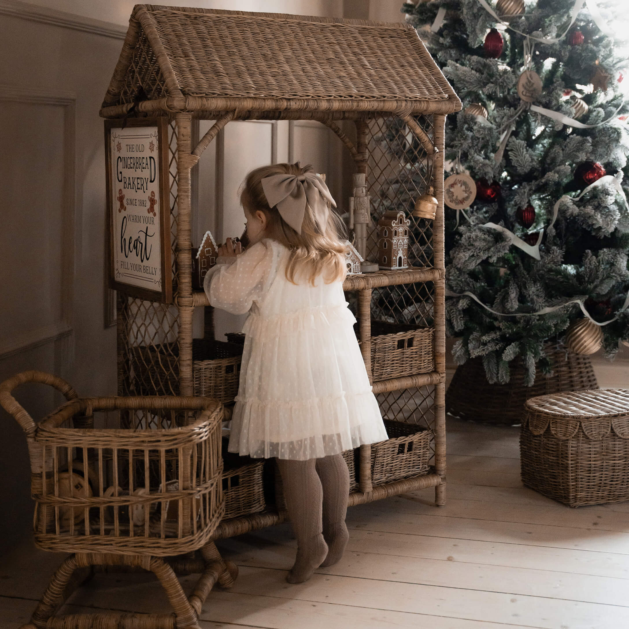 Niña con vestido blanco junto a estantería de mimbre con casas de jengibre cerca de árbol de Navidad decorado