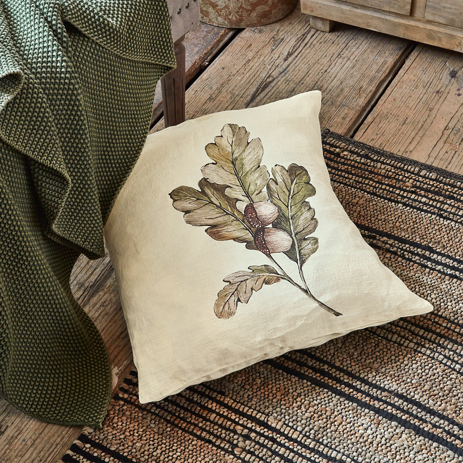 Cojín de lino beige con estampado de hojas y bellotas sobre suelo de madera junto a manta verde