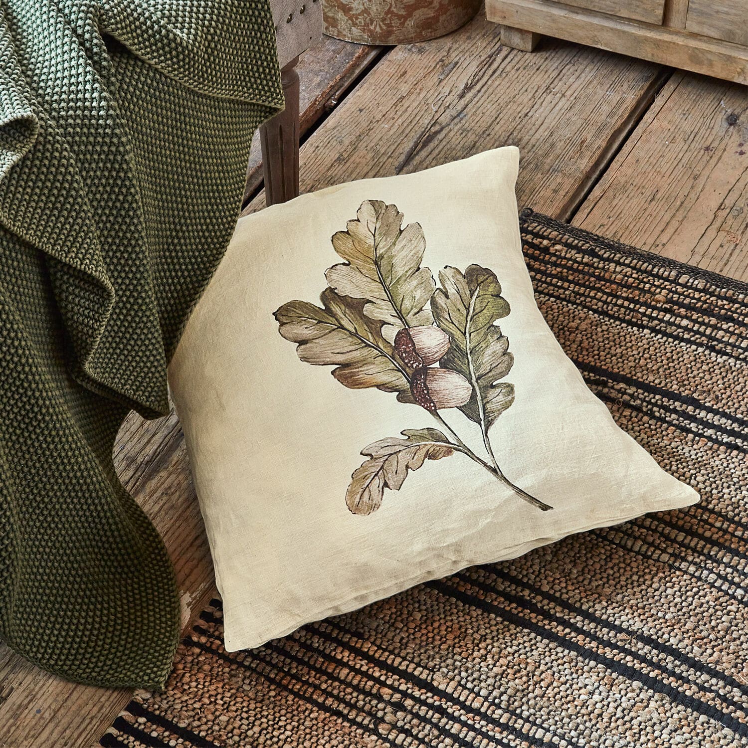 Funda de cojín Vibius Cojín de lino beige con estampado de hojas y bellotas sobre suelo de madera junto a manta verde