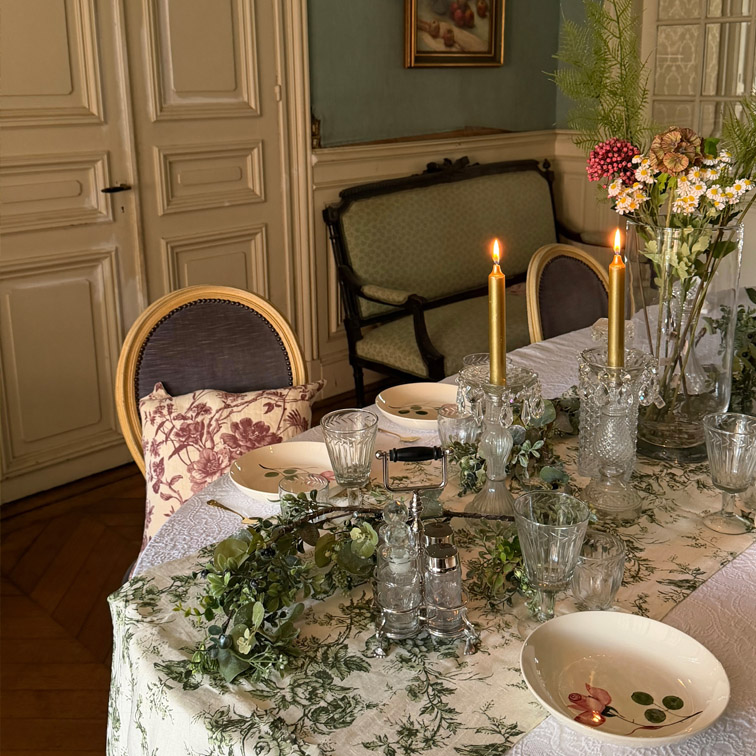 Mesa elegante con mantel blanco de motivos verdes, velas encendidas y decoración floral en interior clásico