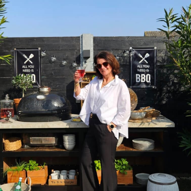 Mujer con camisa blanca y pantalones negros junto a una barbacoa exterior con cartel de BBQ sobre pared negra