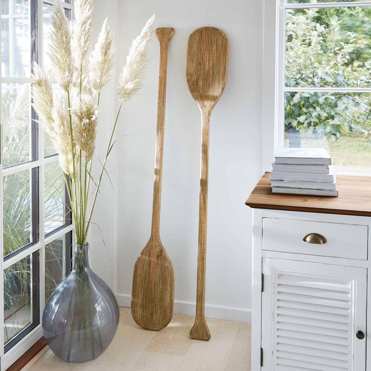 Dos remos decorativos de madera en color natural apoyados en pared blanca en interior luminoso