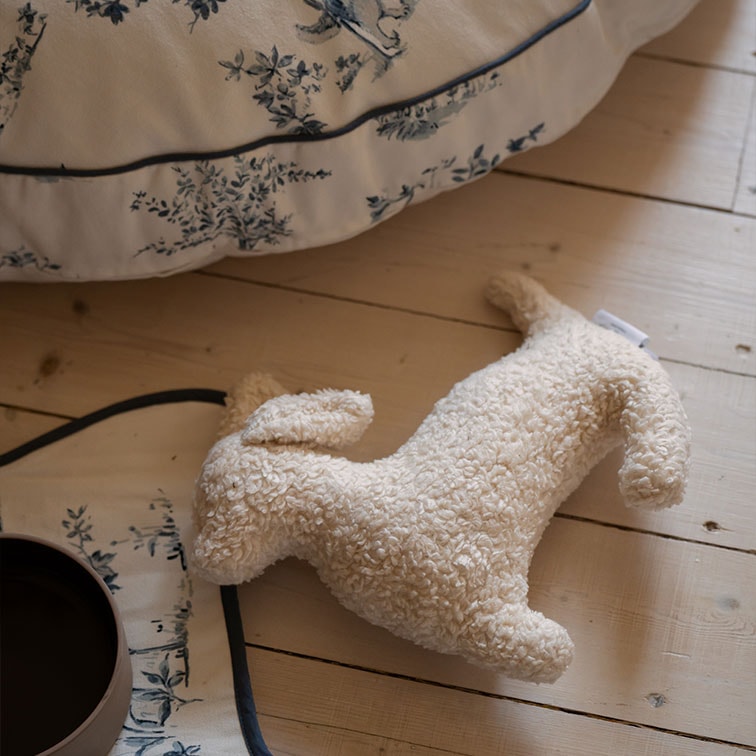 Juguete de peluche suave con forma de perro color crema sobre suelo de madera clara