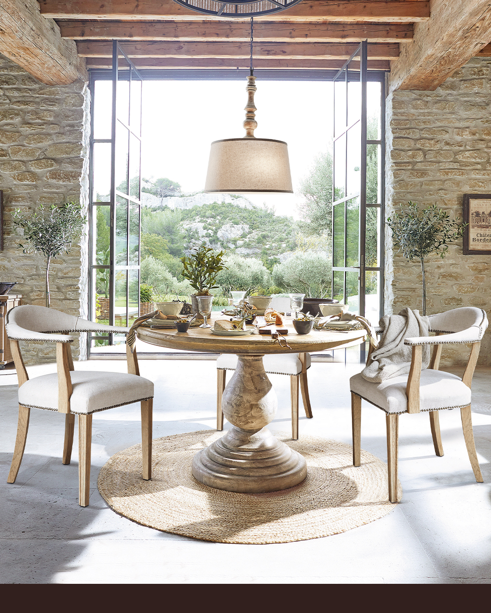 Comedor con mesa redonda de madera y sillas tapizadas beige frente a pared de piedra y grandes ventanas