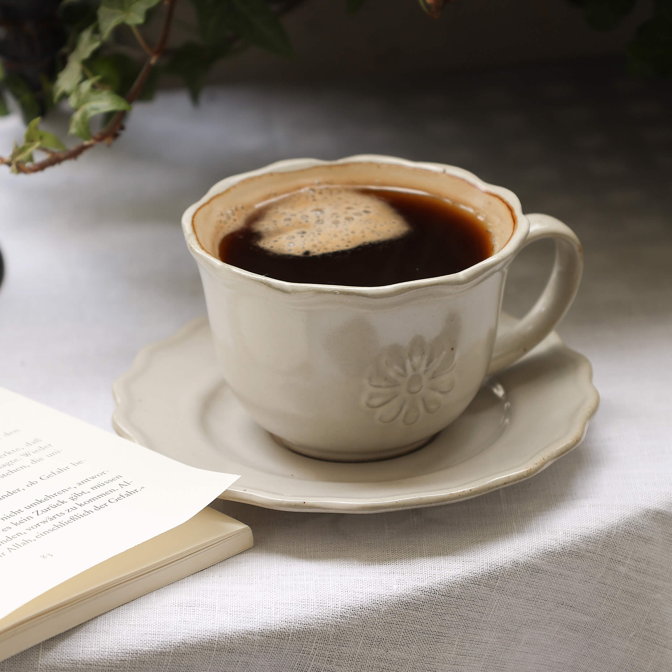 Taza de cerámica beige con café sobre plato en mantel blanco junto a libro abierto