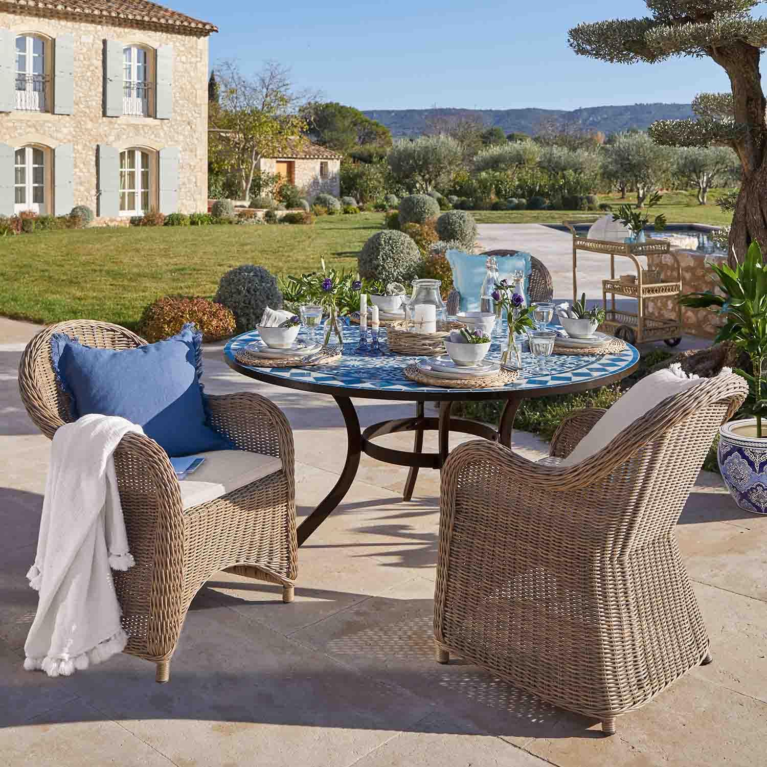Mesa redonda con mosaico azul y sillas de ratán beige en terraza soleada de jardín