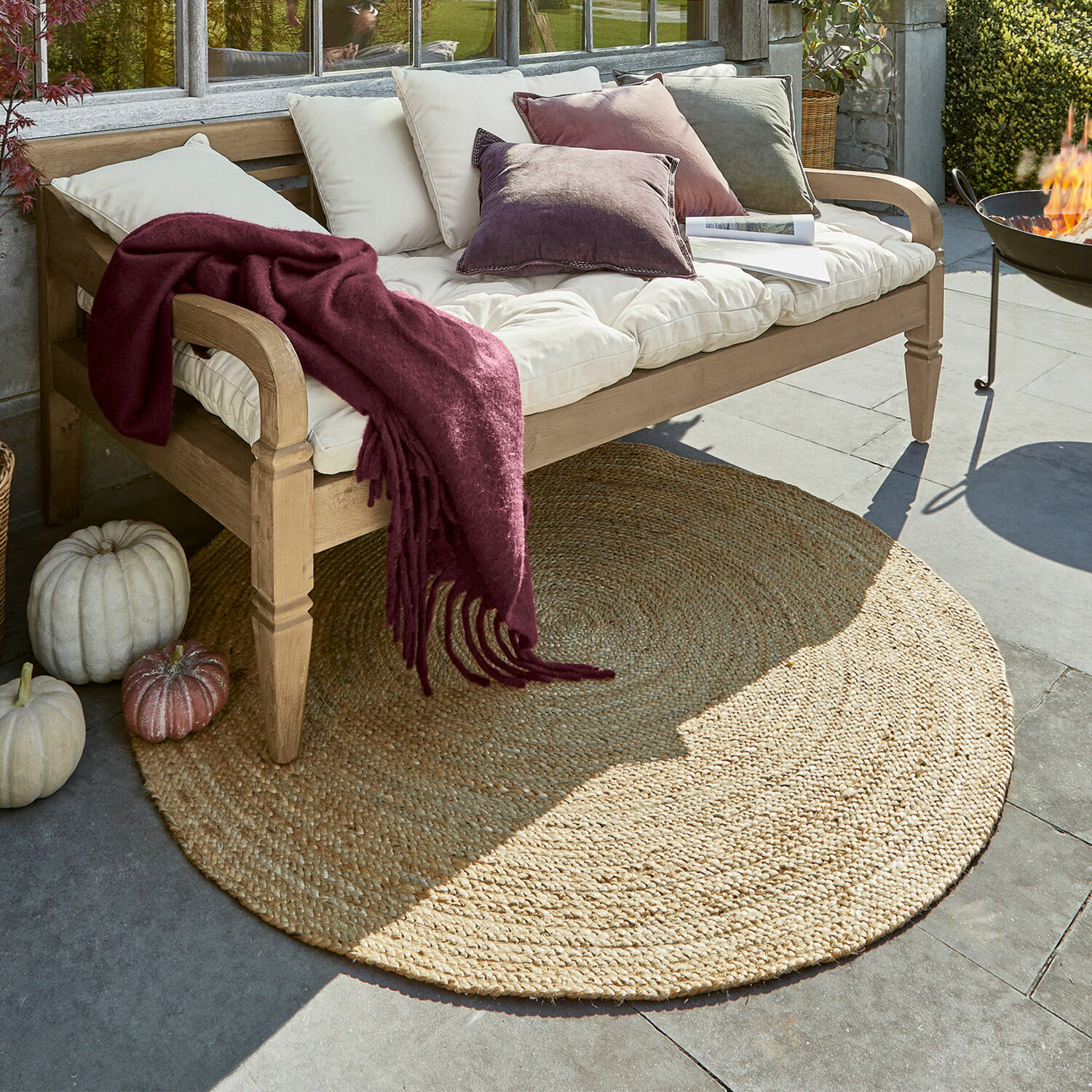 Alfombra redonda de yute color natural bajo banco de madera con cojines y manta burdeos