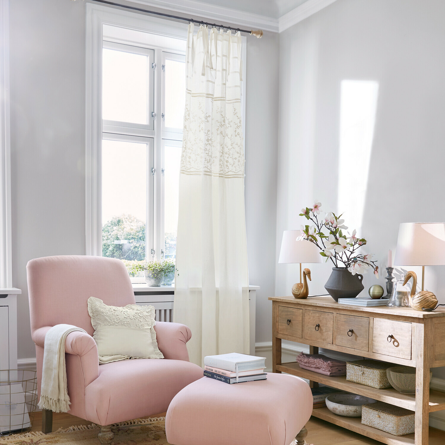 Cortina Lunérie Sillón rosa con reposapiés y cojín crema en salón luminoso con cómoda de madera y decoraciones