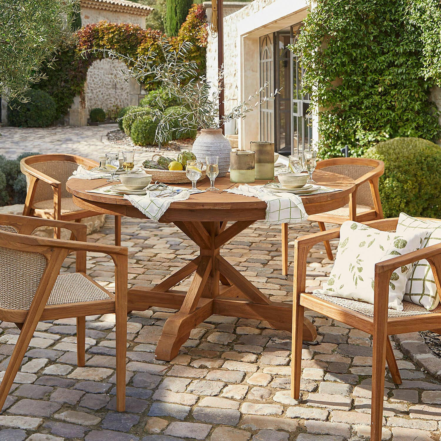 Mesa Wendreau Mesa redonda y sillas de teca en patio empedrado con decoración y vajilla para comedor exterior