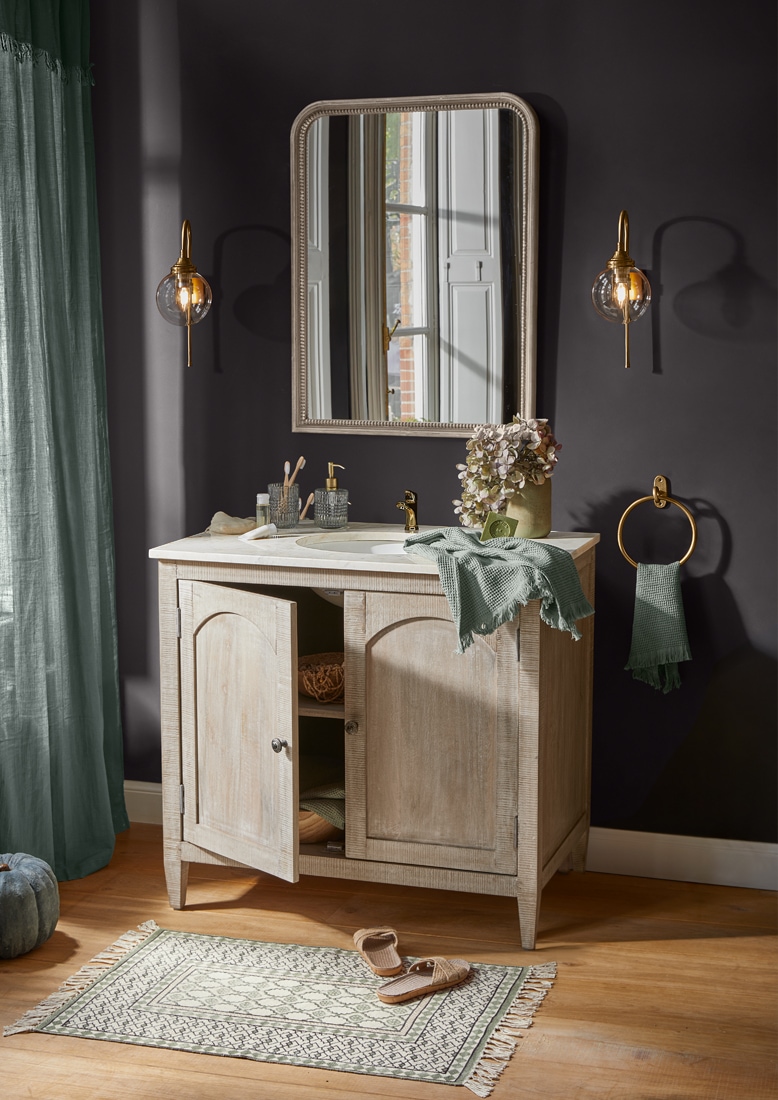 Mueble de baño de madera clara con espejo, accesorios dorados y toalla verde, estilo vintage