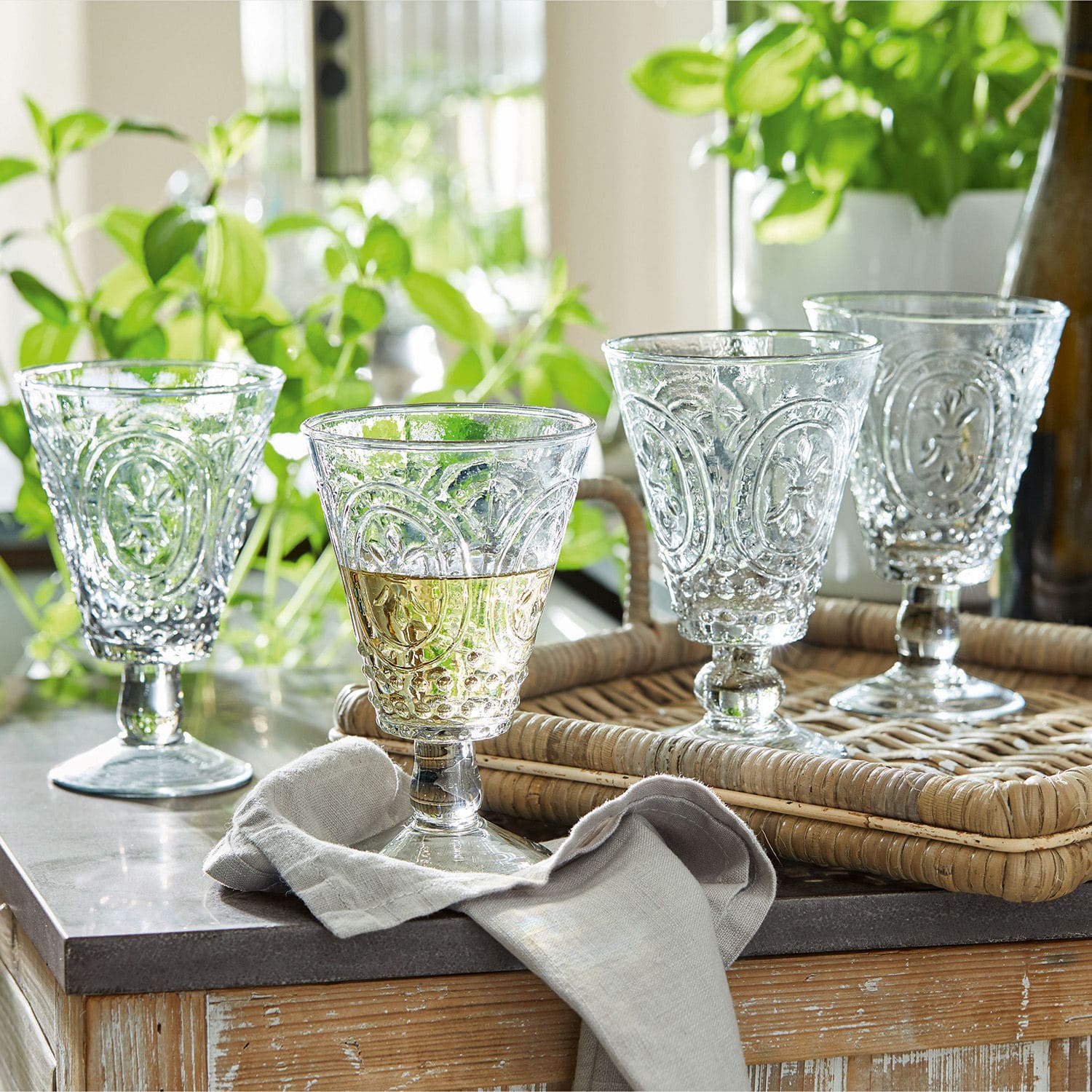 Set de 4 copas de vino Sarton Conjunto de cuatro vasos transparentes con diseño decorativo sobre bandeja de ratán en mesa