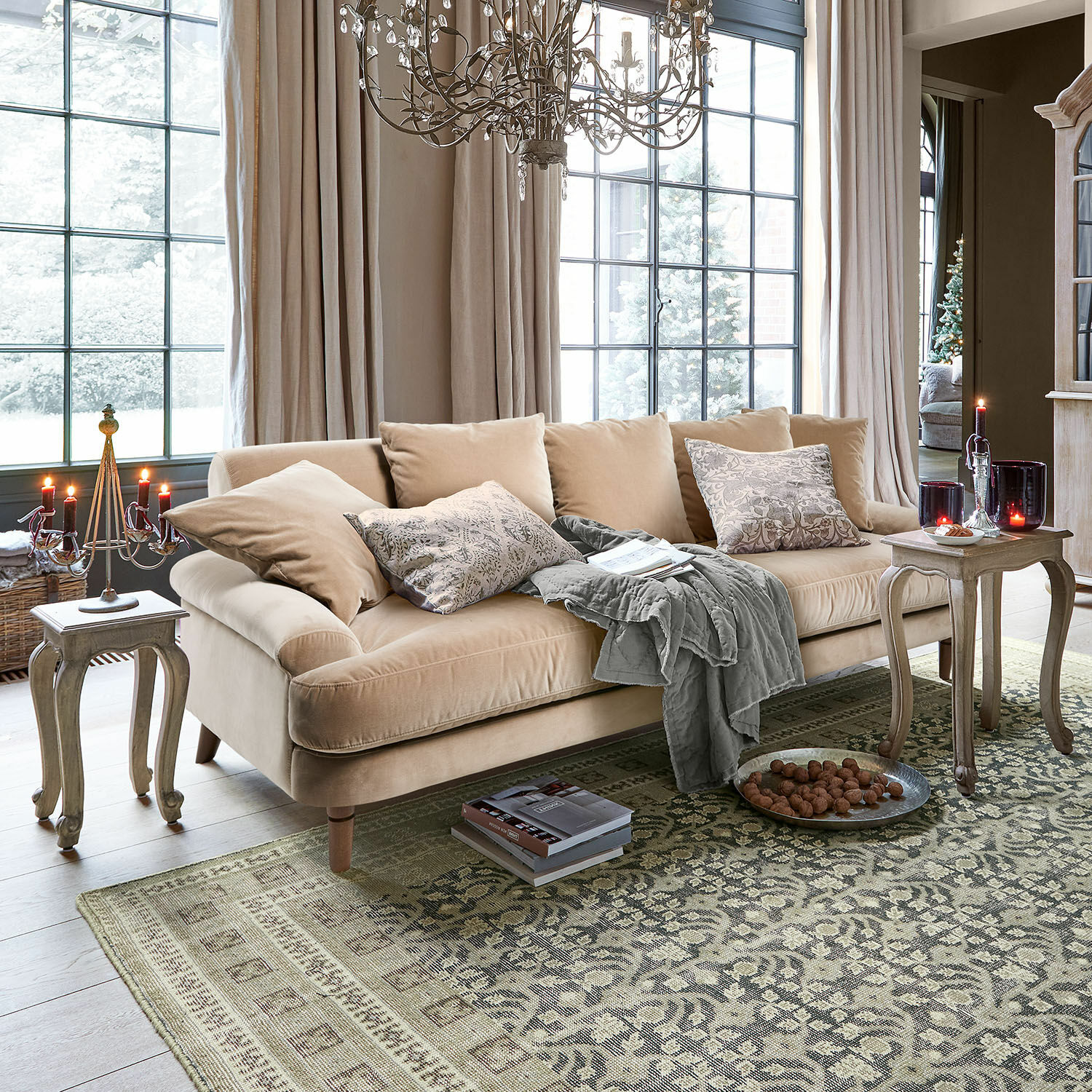 Sofá Rivermere Sofá beige de terciopelo con cojines y manta gris en sala luminosa con alfombra y velas