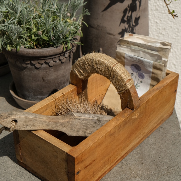 Caja de madera con asa envuelta en cuerda, contiene cepillo y sobres de semillas, materiales naturales